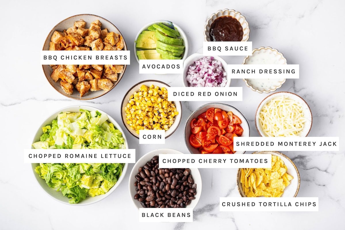BBQ Chicken Salad ingredients: BBQ chicken breasts, avocados, BBQ sauce, ranch dressing, diced red onion, corn, chopped romaine lettuce, chopped cherry tomatoes, shredded Monterey Jack, black beans, crushed tortilla chips.