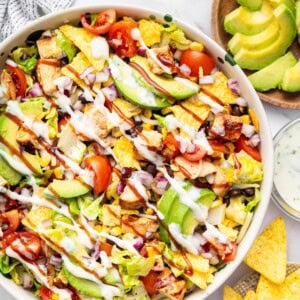 A BBQ chicken salad with avocado slices, BBQ chicken, corn, tortilla chips, tomatoes, lettuce, black beans all topped with a drizzle of BBQ sauce and ranch dressing.
