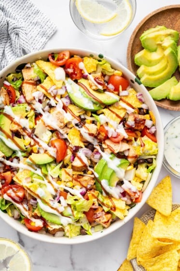 A BBQ chicken salad with avocado slices, BBQ chicken, corn, tortilla chips, tomatoes, lettuce, black beans all topped with a drizzle of BBQ sauce and ranch dressing.