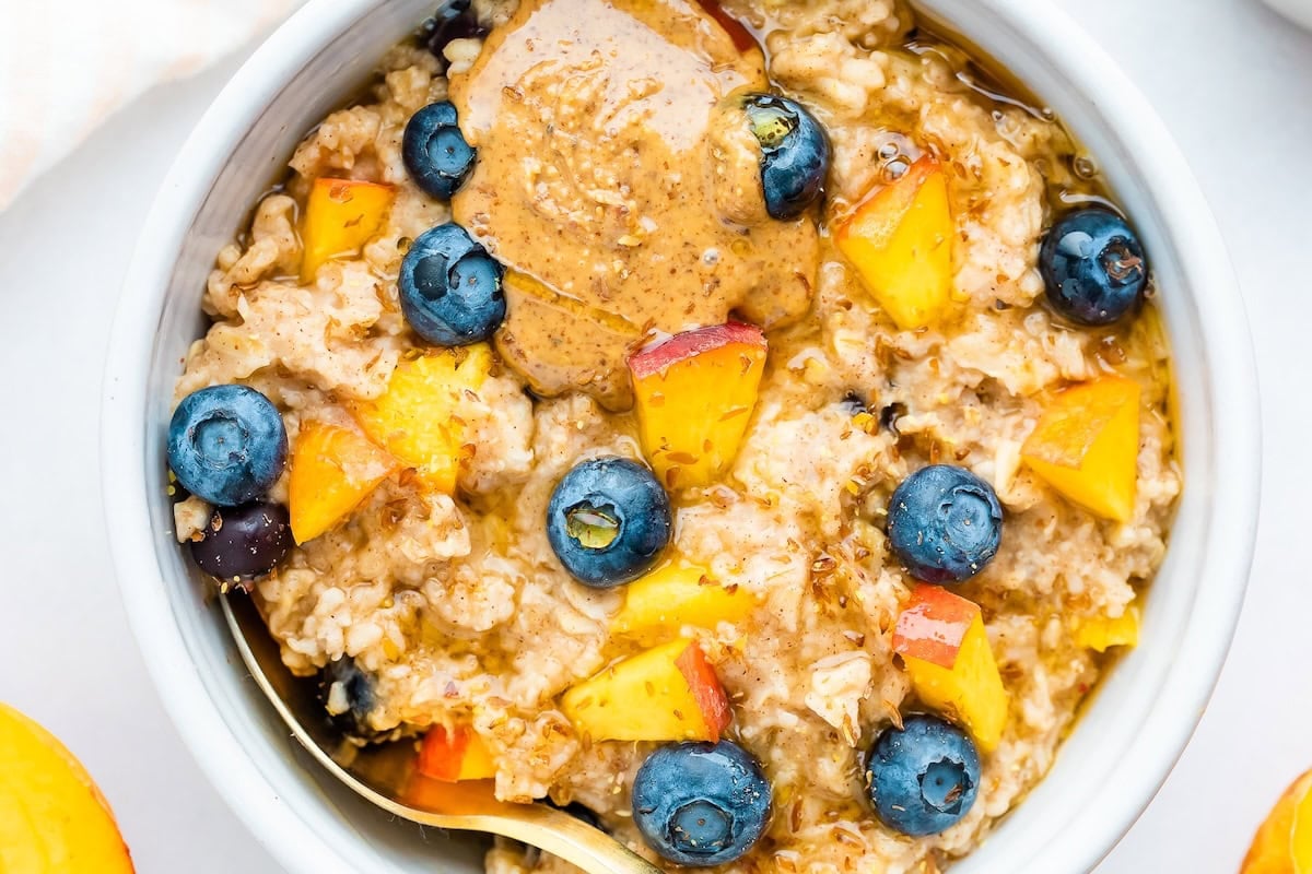 Blueberry Peach Oatmeal - Loaded with fiber, healthy fat and protein this delicious bowl of oats will keep you feeling full and energized all morning. Overhead close up of blueberry peach oatmeal in a white bowl with blueberries and fresh chopped peaches on top with a little almond butter on top as well.