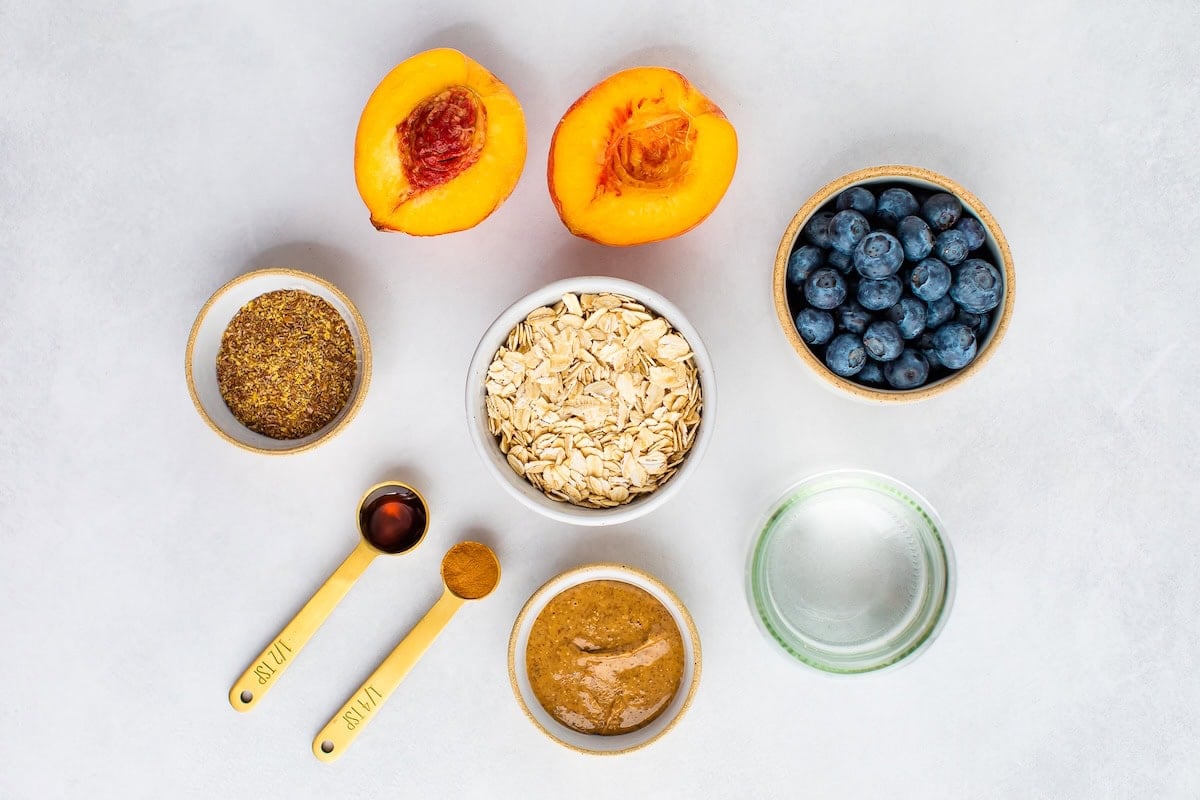 Ingredients measured out to make blueberry peach oatmeal: blueberries, water, peaches, oats, almond butter, cinnamon, vanilla and flaxseed.