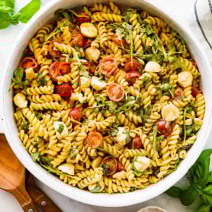 A serving bowl filled with caprese pasta salad. There are serving spoons next to the bowl.
