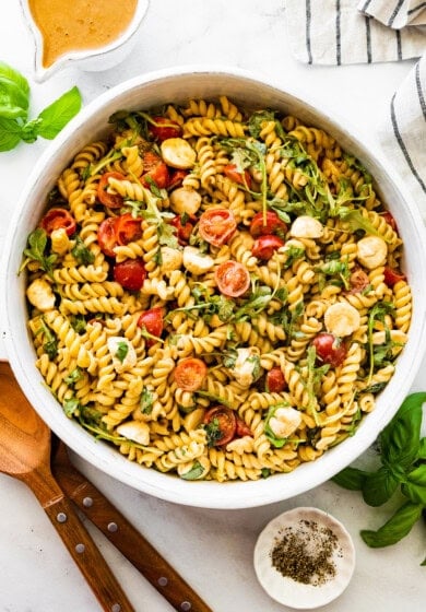 A serving bowl filled with caprese pasta salad. There are serving spoons next to the bowl.