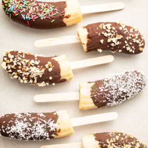 Frozen banana halves on wooden sticks, dipped in chocolate and topped with various toppings including rainbow sprinkles, chopped nuts, and shredded coconut.