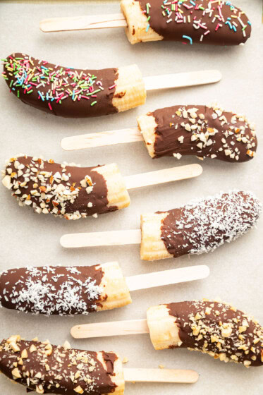 Frozen banana halves on wooden sticks, dipped in chocolate and topped with various toppings including rainbow sprinkles, chopped nuts, and shredded coconut.