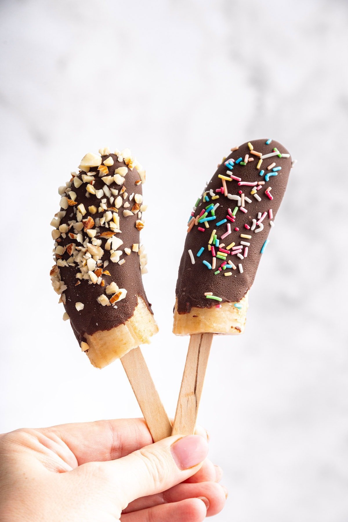 Hand holding two chocolate-dipped banana pops on sticks, one topped with chopped nuts and the other with colorful rainbow sprinkles.