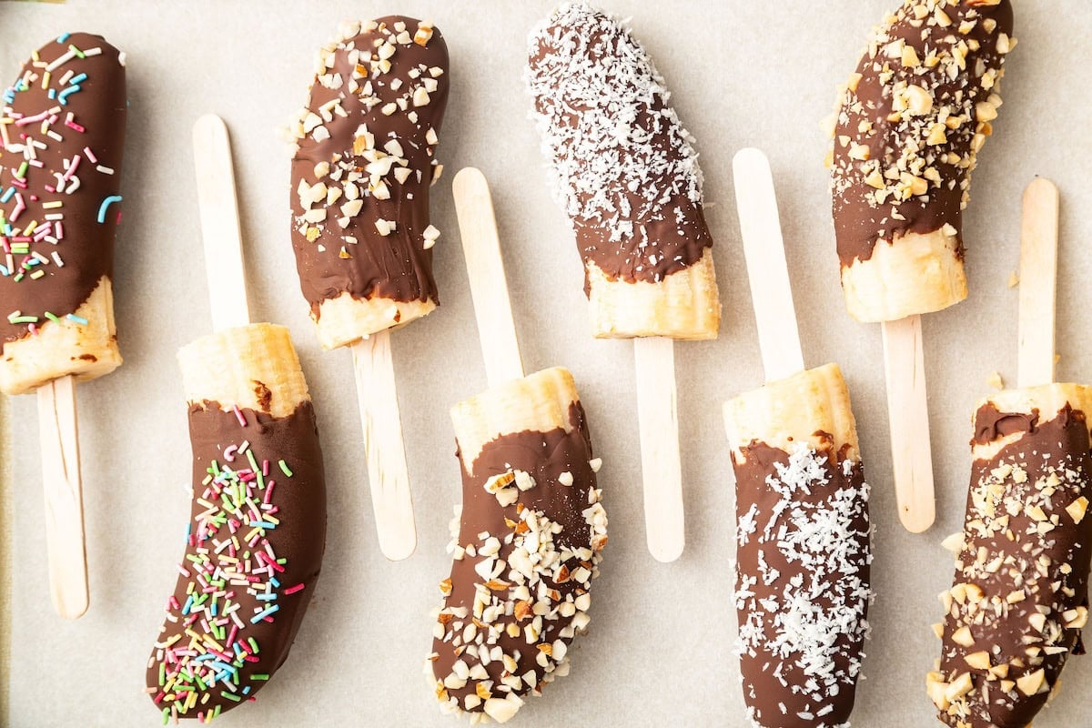 Frozen banana halves on wooden sticks, dipped in chocolate and topped with various toppings including rainbow sprinkles, chopped nuts, and shredded coconut.