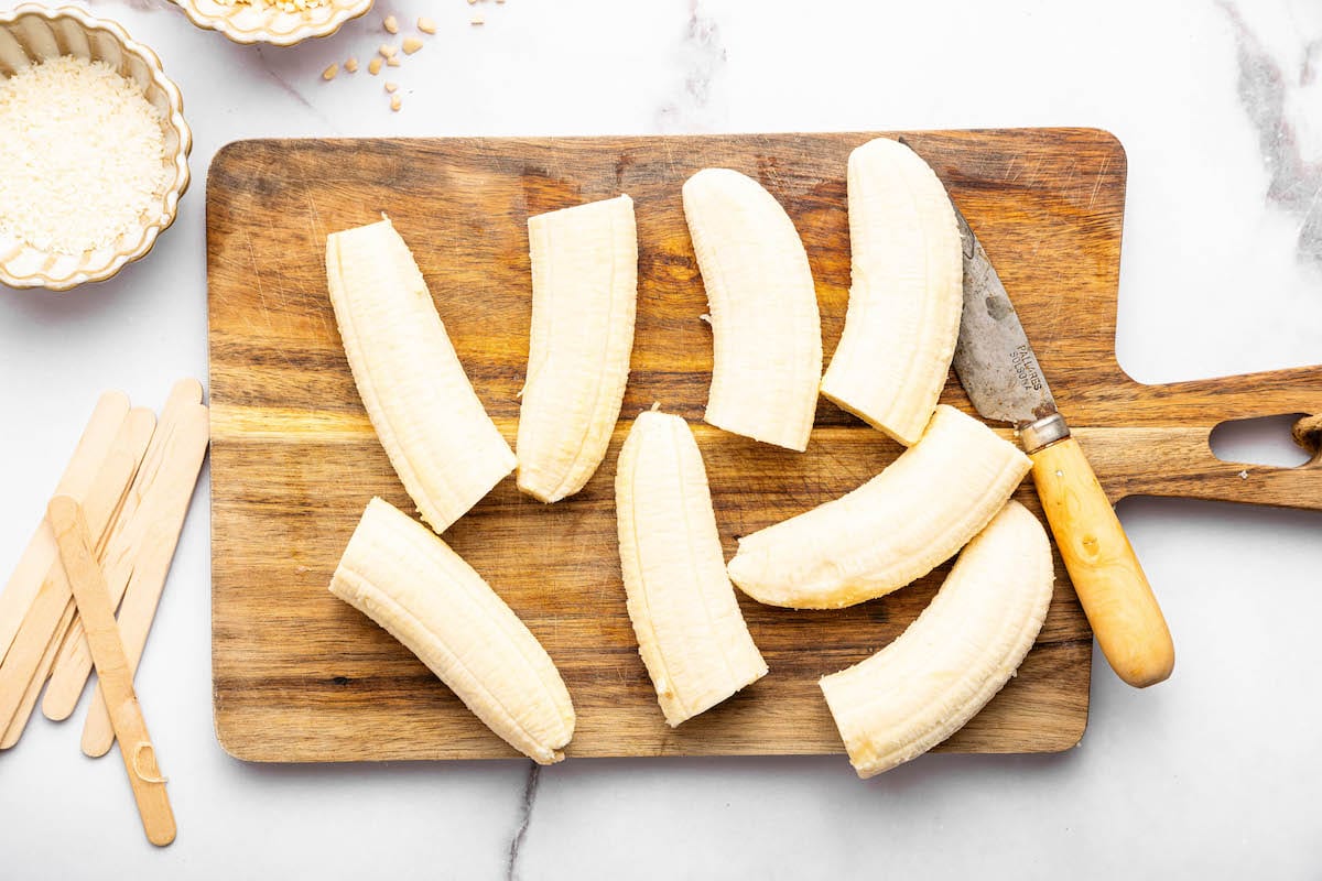 Eight banana halves placed on a wooden cutting board next to a paring knife, with wooden popsicle sticks next to the cutting board.