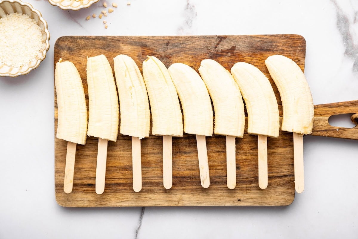 Eight halved bananas on wooden sticks lined up on a wooden cutting board, ready for dipping.