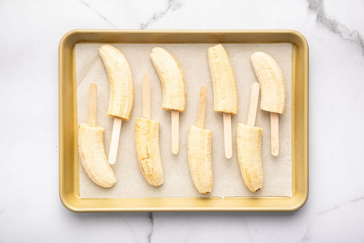 Eight banana halves on wooden sticks arranged on a parchment-lined baking tray, prepared for freezing or dipping, set on a white marble surface.