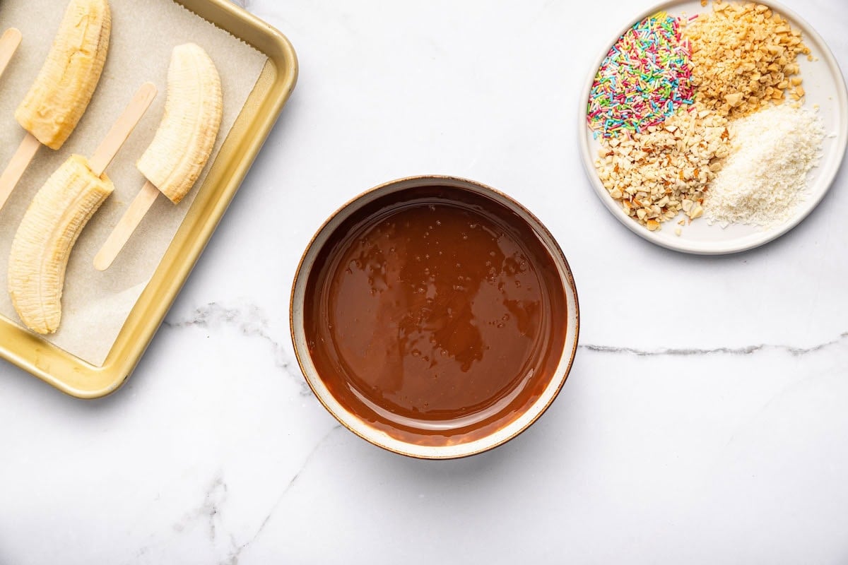 Bowl of melted chocolate, a plate with colorful sprinkles, chopped nuts, and shredded coconut, and a tray of banana halves on sticks, all set on a white marble surface ready for assembling frozen banana pops.
