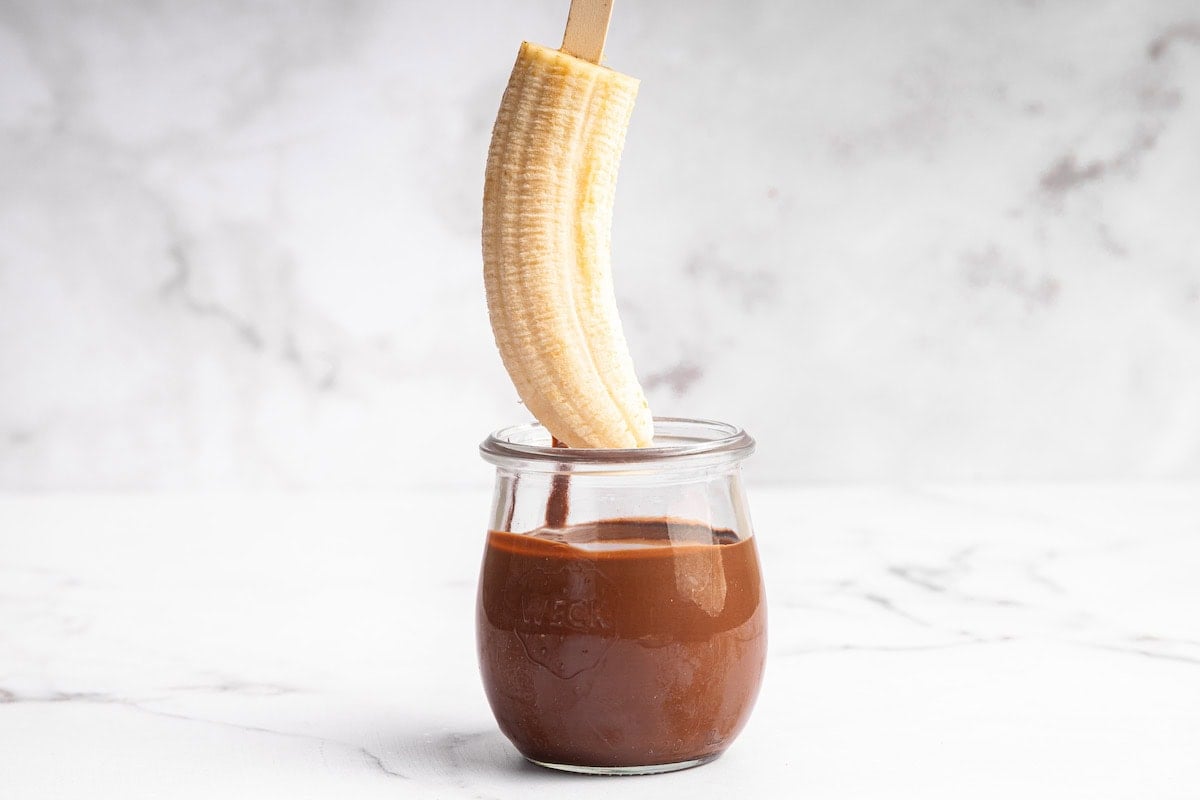 Banana half on a wooden stick being dipped into a small glass jar of melted chocolate, set on a white marble surface.