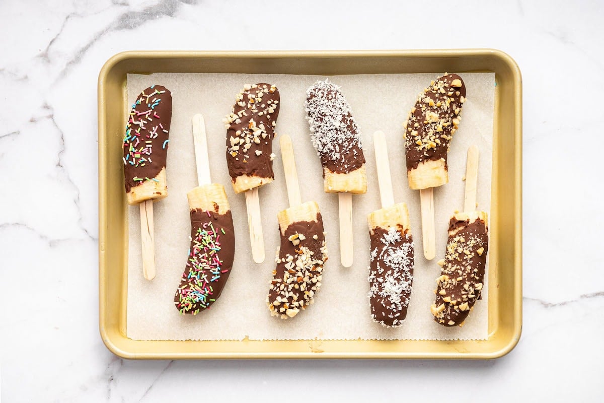 Tray of chocolate-dipped banana halves on sticks, topped with rainbow sprinkles, chopped nuts, and shredded coconut, arranged on parchment paper.