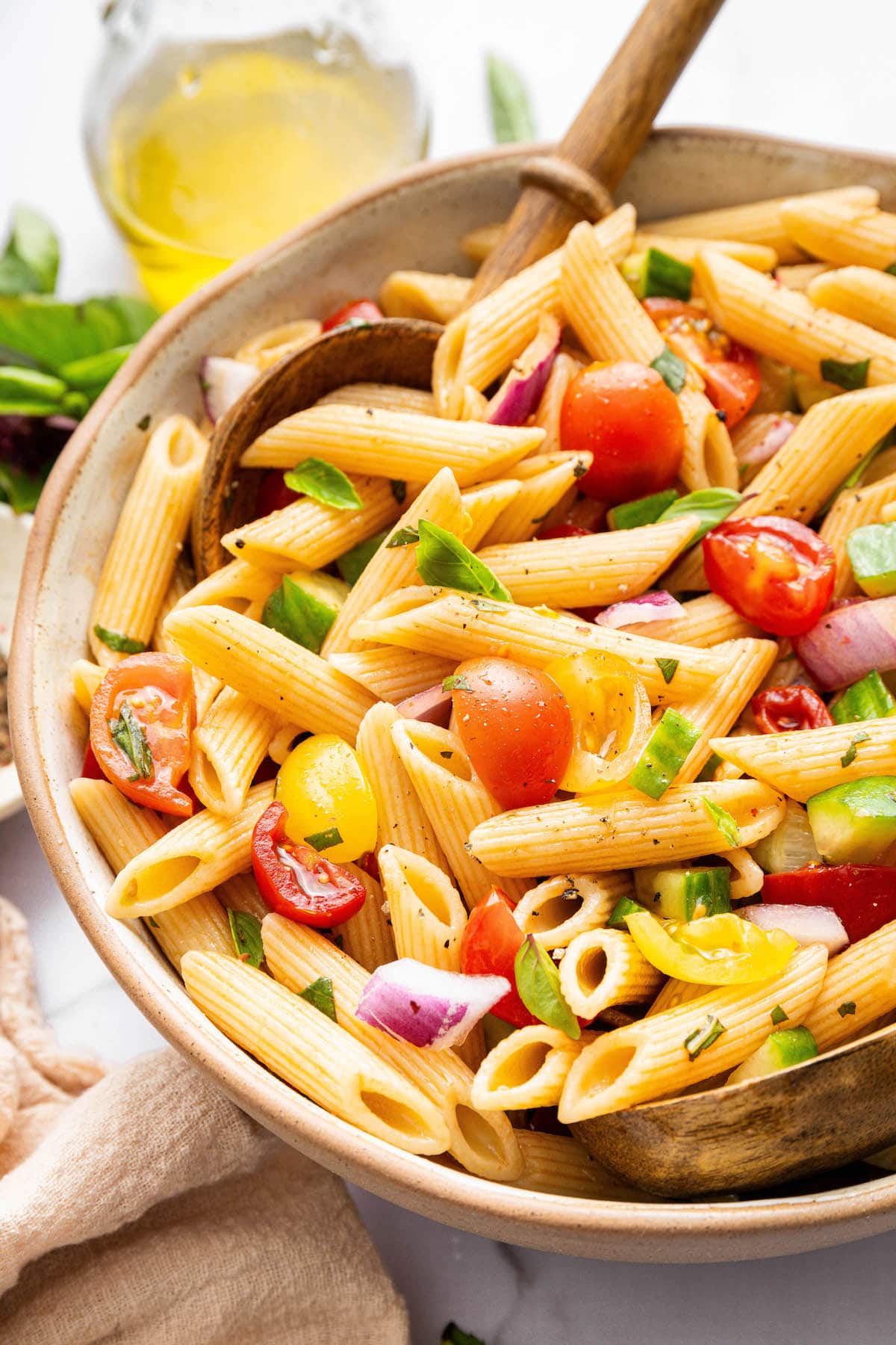 A pasta salad in serving bowl with cherry tomatoes, cucumber, red onion and fresh basil. There are two wooden serving spoons in the bowl.