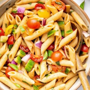 A pasta salad in serving bowl with cherry tomatoes, cucumber, red onion and fresh basil. There is a wooden serving spoon in the bowl.