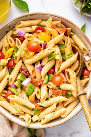 A pasta salad in serving bowl with cherry tomatoes, cucumber, red onion and fresh basil. There is a wooden serving spoon in the bowl.