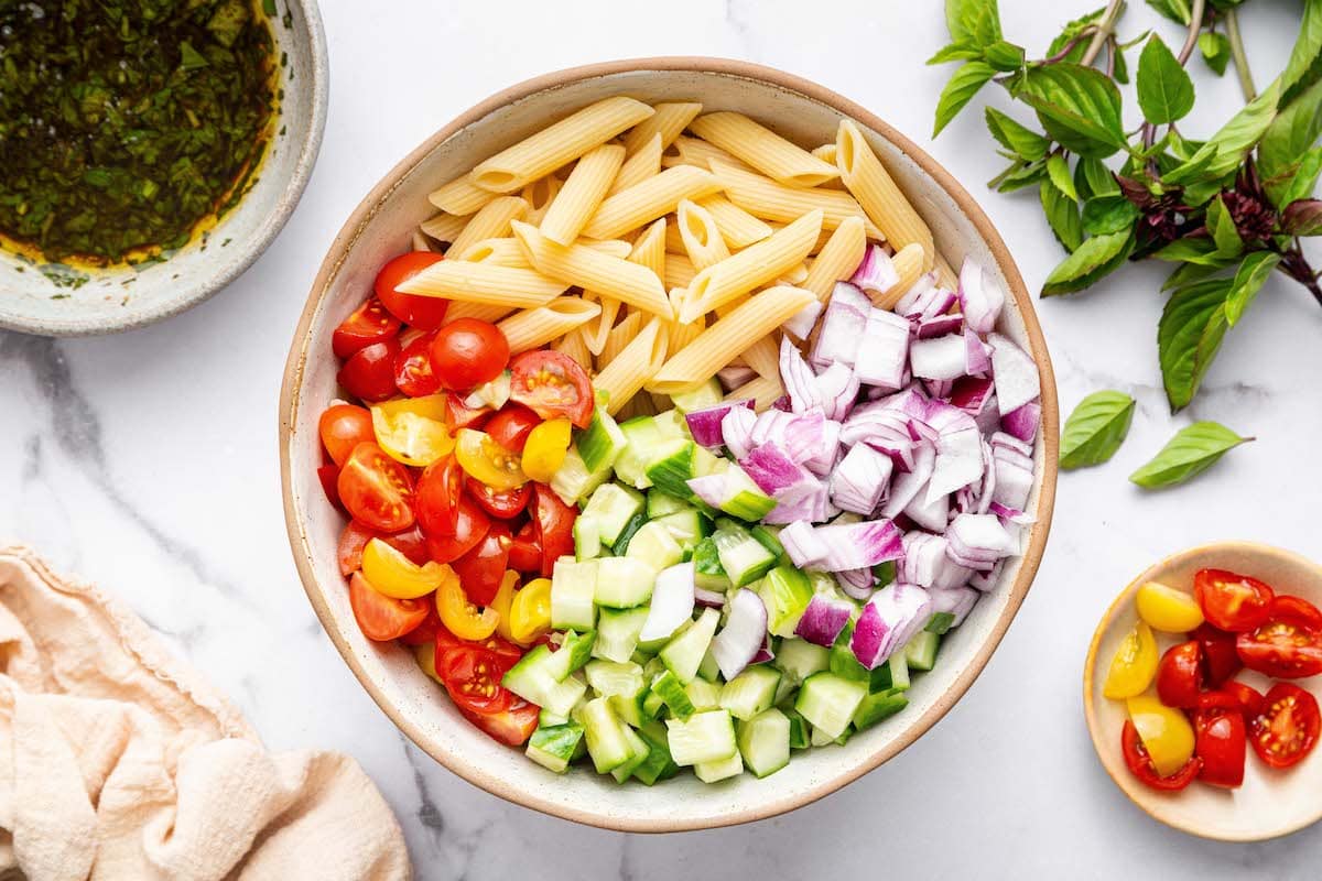 Ingredients for the healthy pasta salad in a large bowl: cooked pasta, red onion, cherry tomatoes and cucumber.