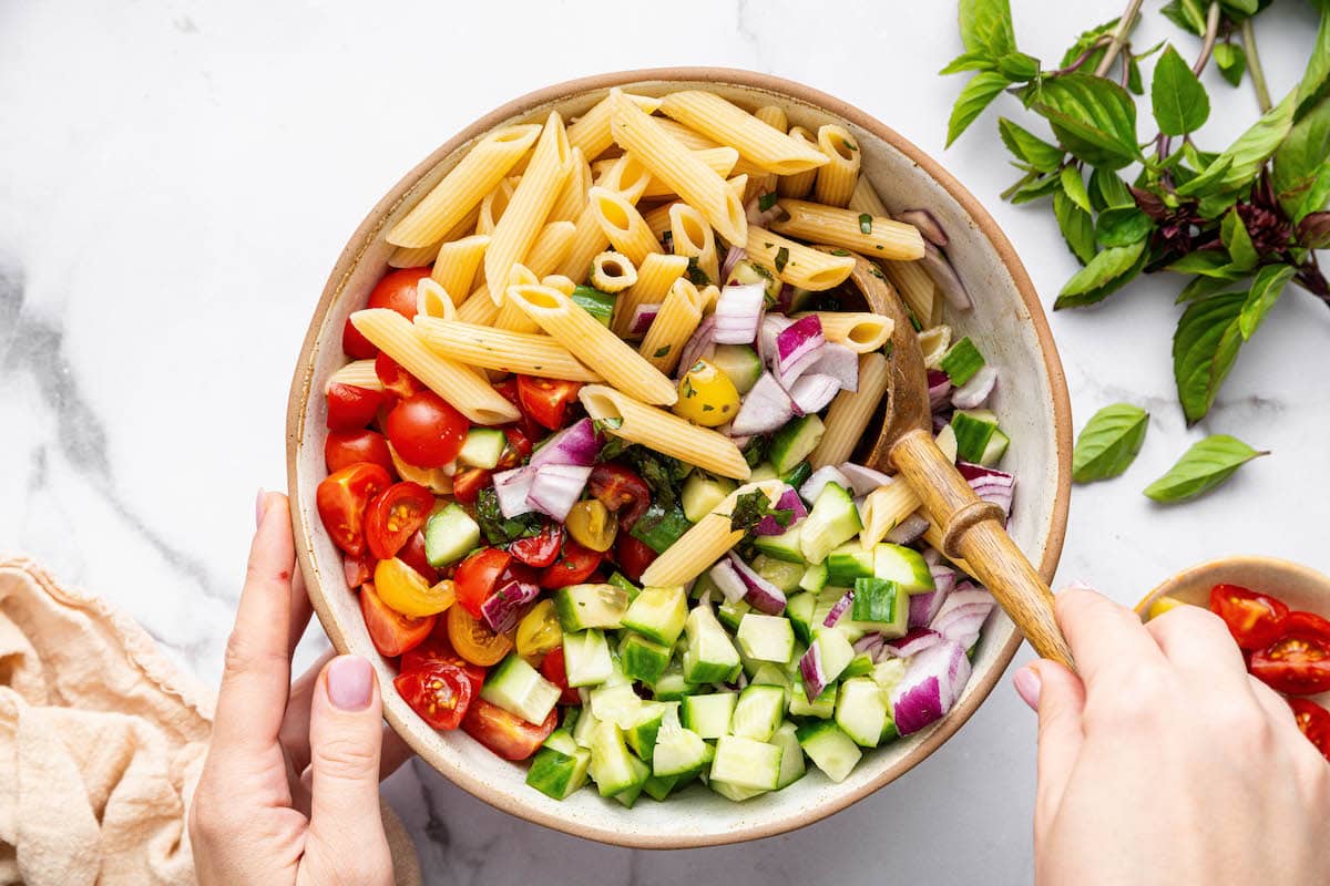 A woman's hand mixing together the healthy pasta salad with a wooden serving spoon.