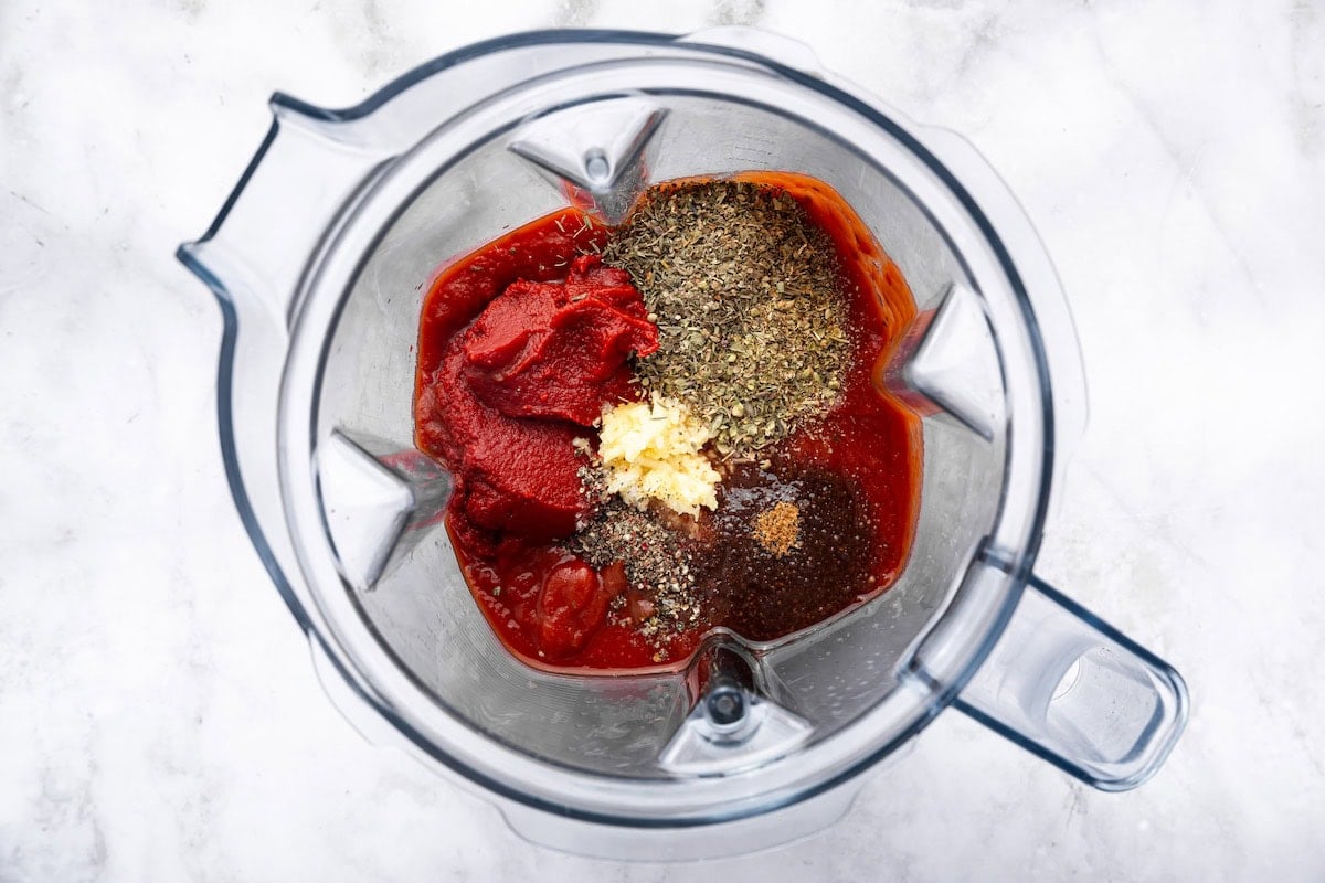 Blender filled with tomato paste, tomato sauce, garlic, and dried herbs before blending pizza sauce.