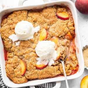 Overhead view of a baked peach cobbler with golden brown topping and two scoops of vanilla ice cream in a white square dish.