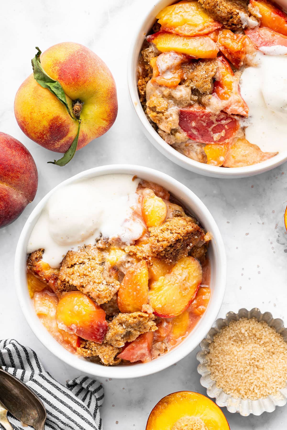 Two white bowls filled with healthy peach cobbler and vanilla ice cream, surrounded by fresh peaches and a bowl of brown sugar.