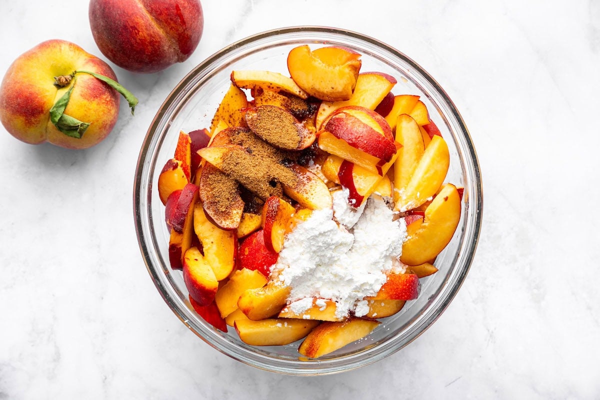Unbaked peach cobbler in a glass bowl with sliced fresh peaches, cinnamon topping, and arrowroot powder ready to be mixed.