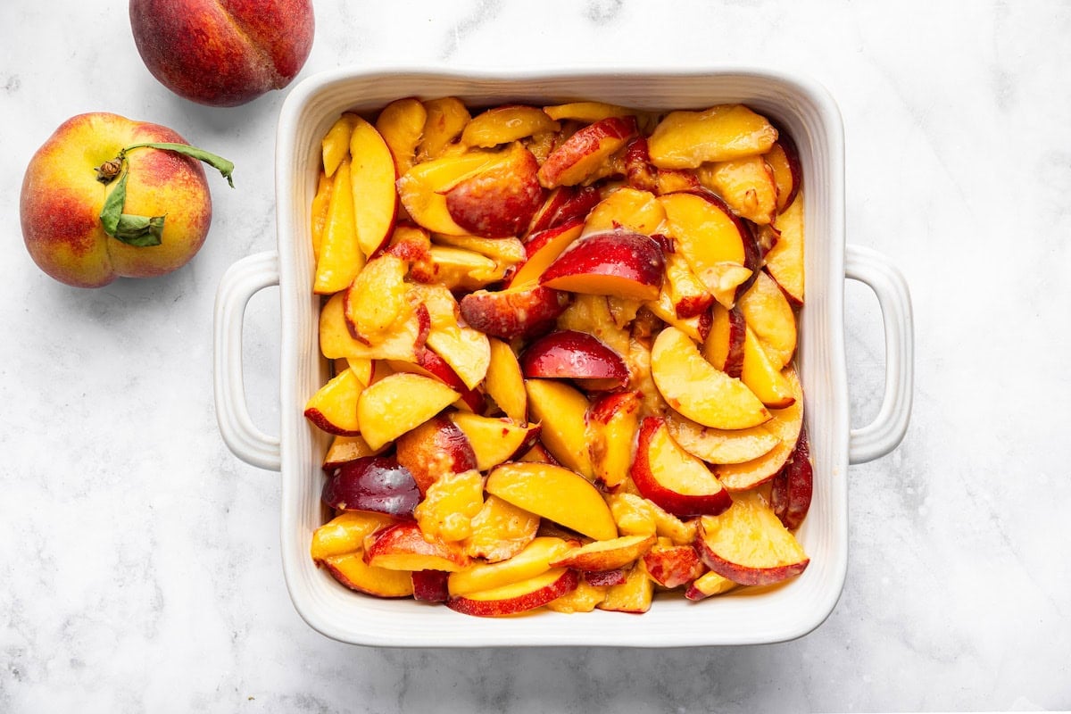 White baking dish filled with sliced fresh peaches coated in arrowroot and lemon juice, ready to be topped and baked.