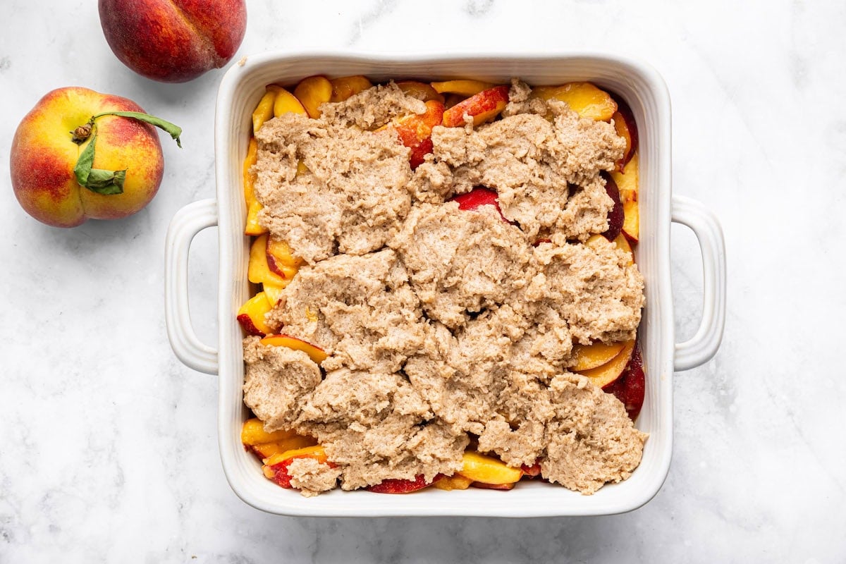 Unbaked peach cobbler in a white baking dish with topping mixture spooned over fresh peach filling.