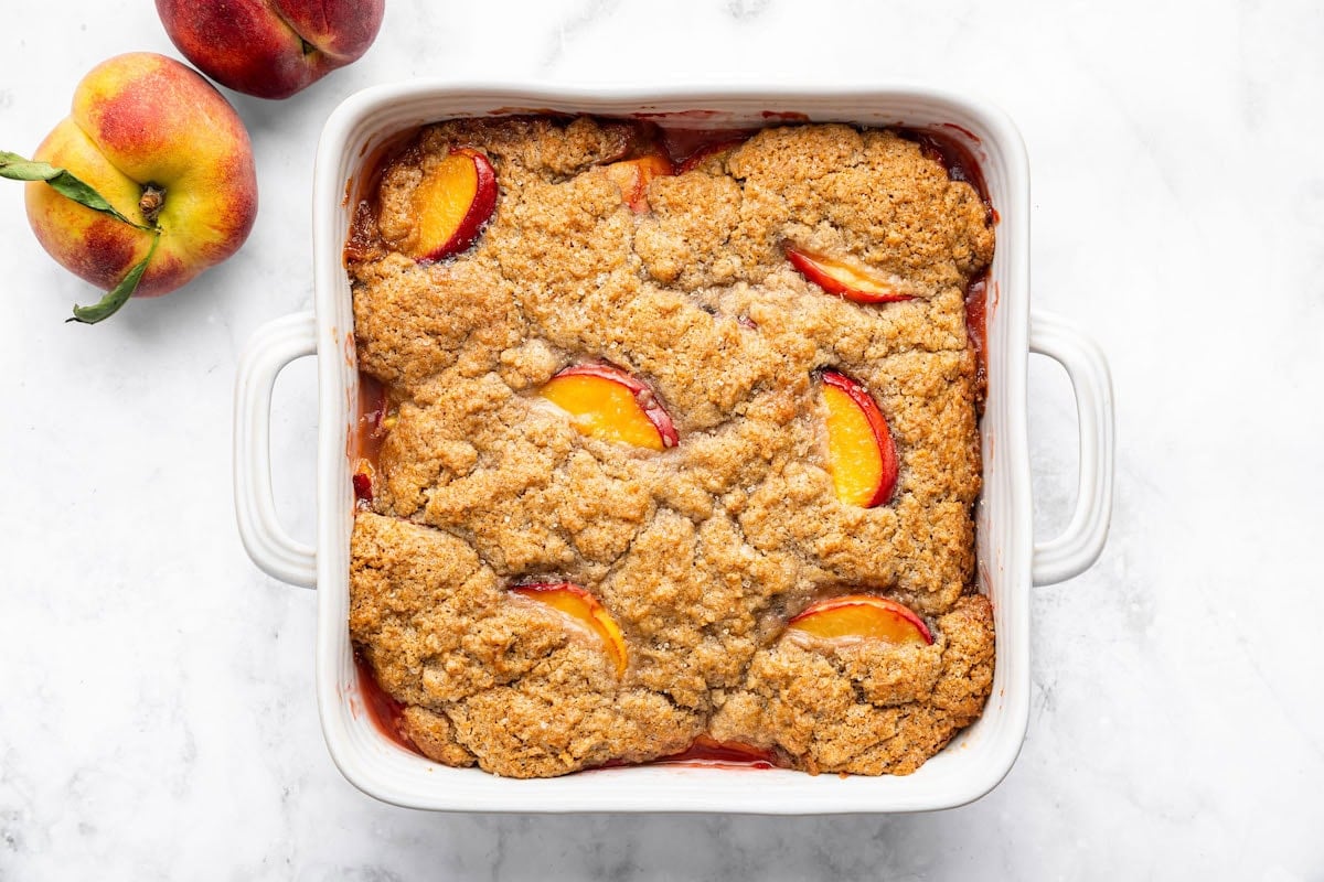 Baked healthy peach cobbler in a white dish with a golden brown, cinnamon-spiced topping and visible peach slices.