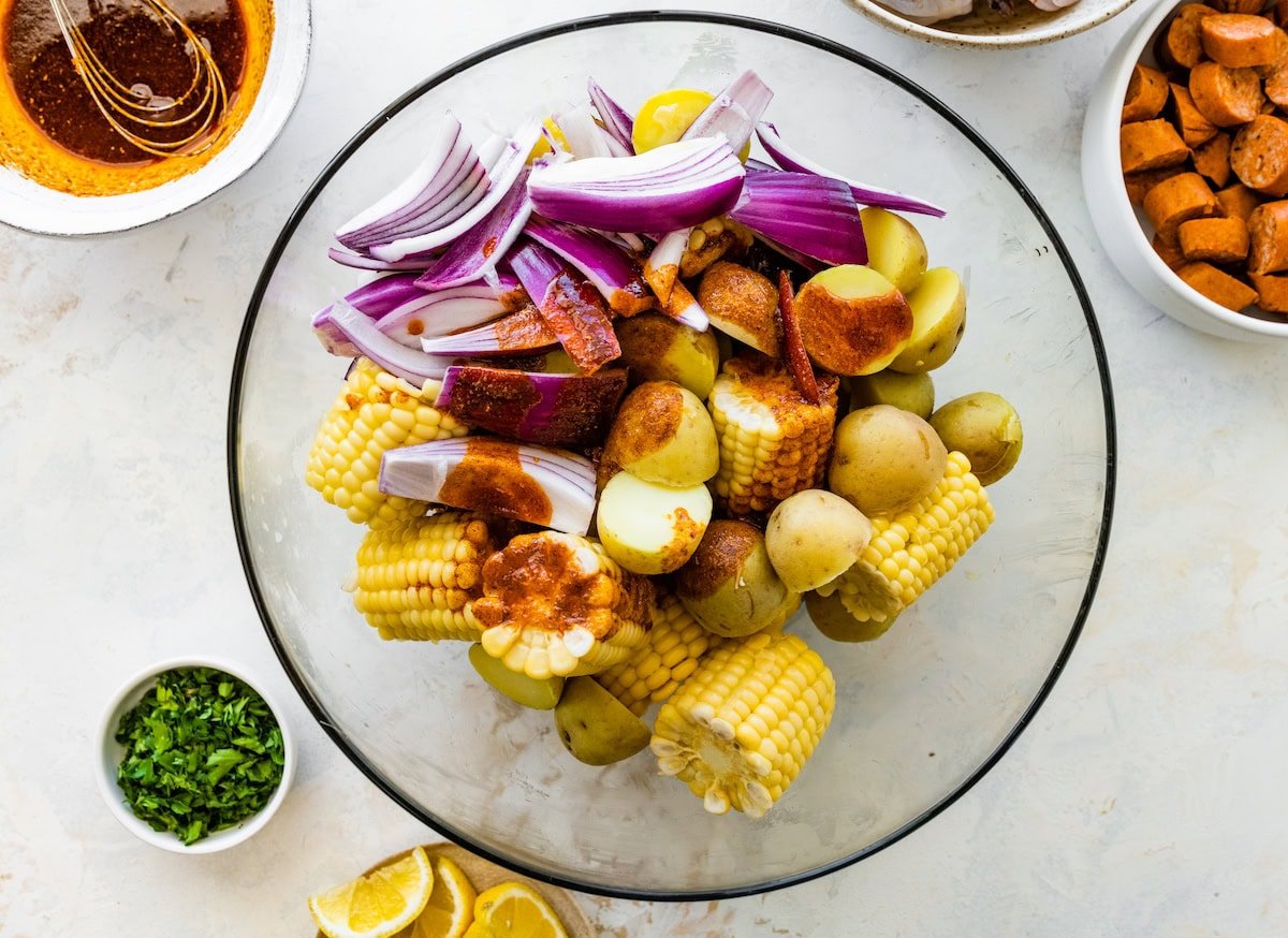 Fresh corn, potatoes, and red onion in a glass bowl with butter mixture poured on top before mixing.