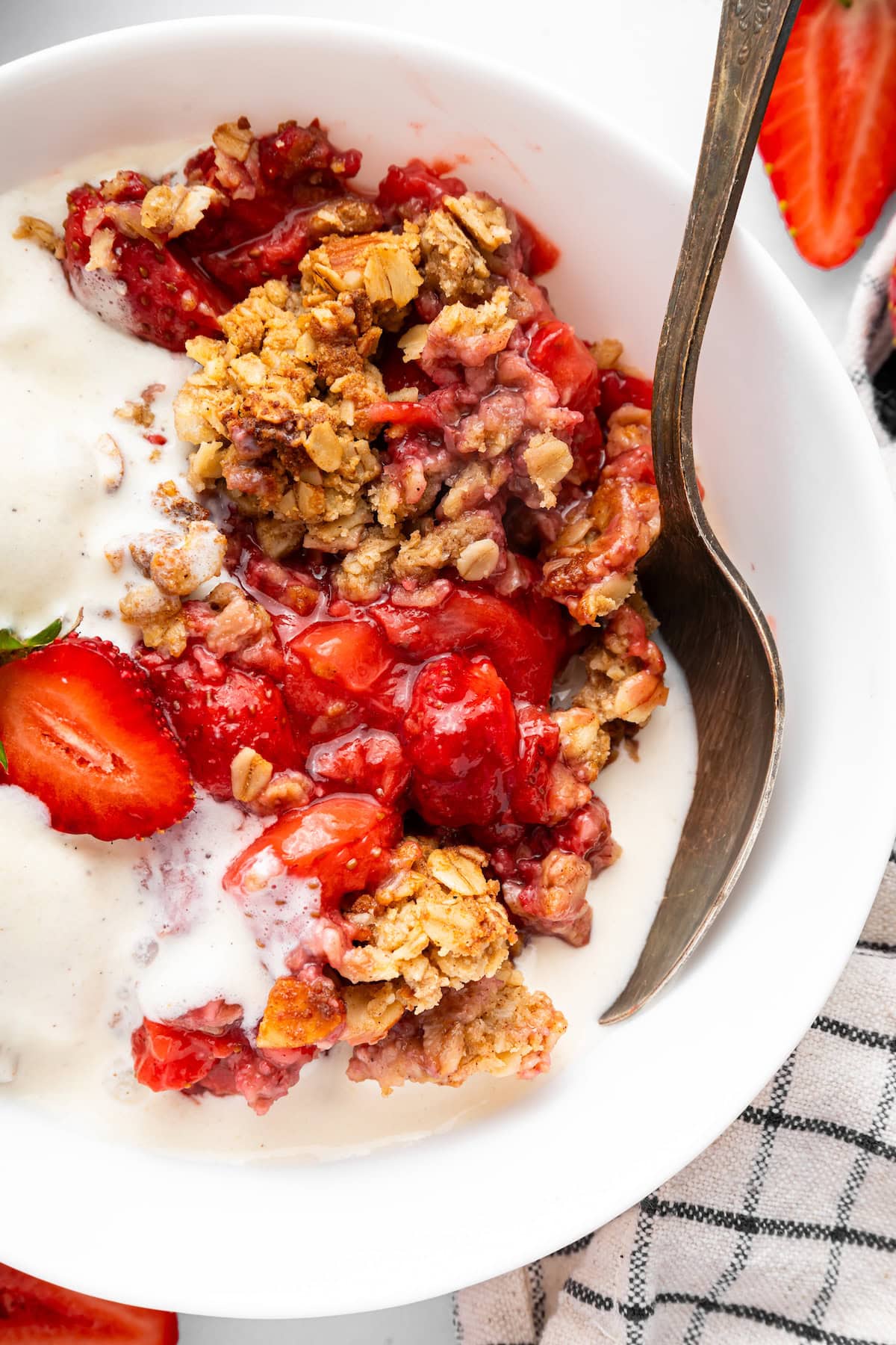 A bowl of strawberry crisp served with vanilla ice cream. There is a silver spoon in the bowl.