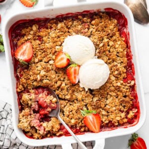 Baked strawberry crisp topped with two scoops of vanilla ice cream and fresh strawberry halves. There is a spoon scooping out some of the crisp.