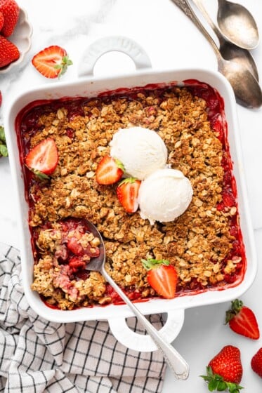 Baked strawberry crisp topped with two scoops of vanilla ice cream and fresh strawberry halves. There is a spoon scooping out some of the crisp.