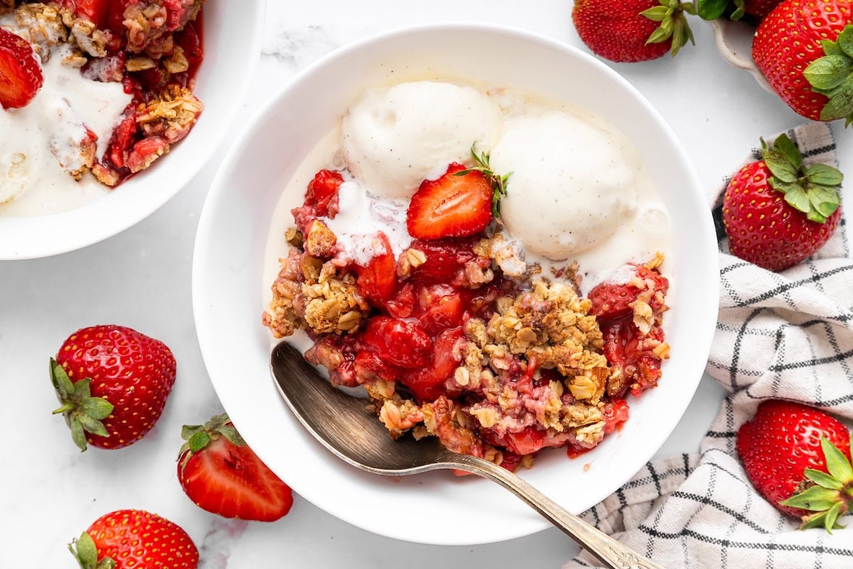 Two bowls of strawberry crisp served with vanilla ice cream. There are fresh strawberries surrounding the bowls.