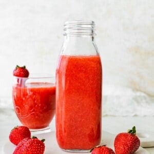 Glass bottle filled with strawberry juice placed on a white surface, with a glass of juice and strawberries beside it.
