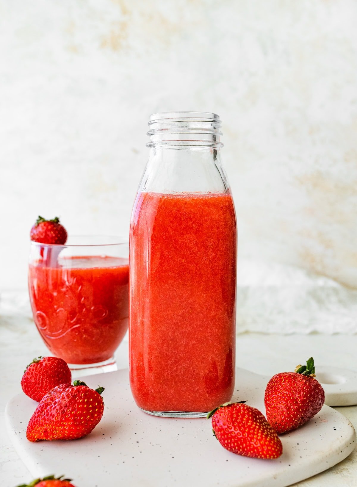 Glass bottle filled with strawberry juice placed on a white surface, with a glass of juice and strawberries beside it.