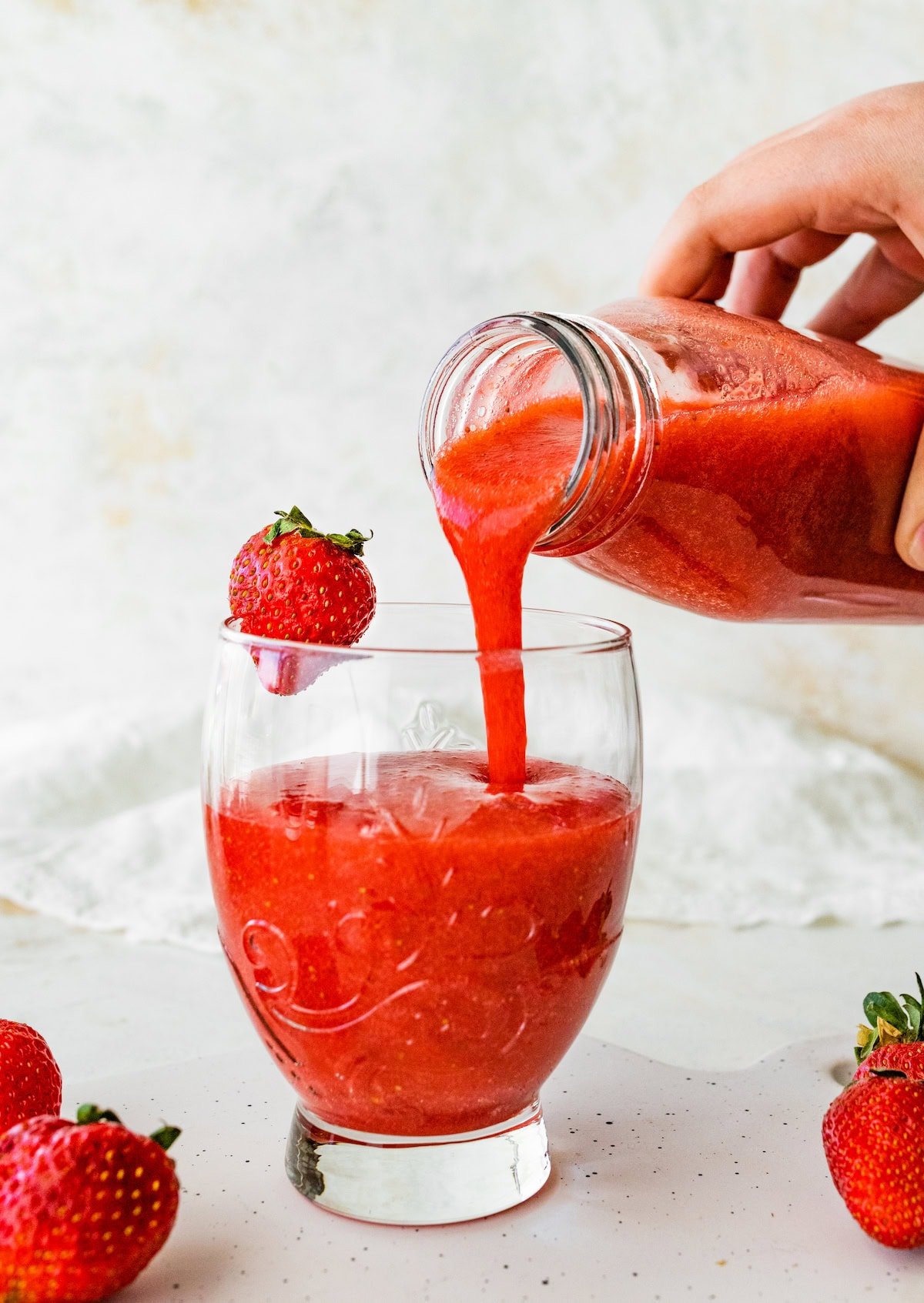 Strawberry juice being poured from a bottle into a glass garnished with a fresh strawberry, on a white speckled surface.
