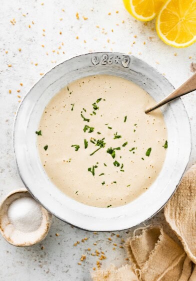 Bowl of creamy tahini sauce topped with fresh chopped parsley, served with a spoon and surrounded by lemon slices and sesame seeds.