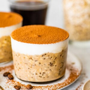 Glass jar of tiramisu overnight oats topped with Greek yogurt and cocoa powder, styled on a plate with coffee beans and spoons.