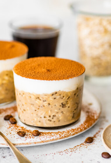 Glass jar of tiramisu overnight oats topped with Greek yogurt and cocoa powder, styled on a plate with coffee beans and spoons.