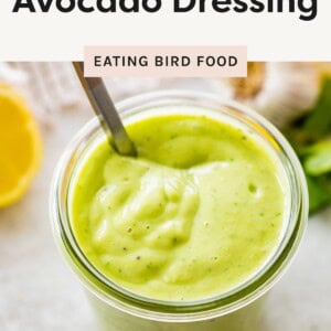 Glass jar filled with thick, creamy avocado dressing with a spoon inside, surrounded by fresh ingredients like lemon and garlic.