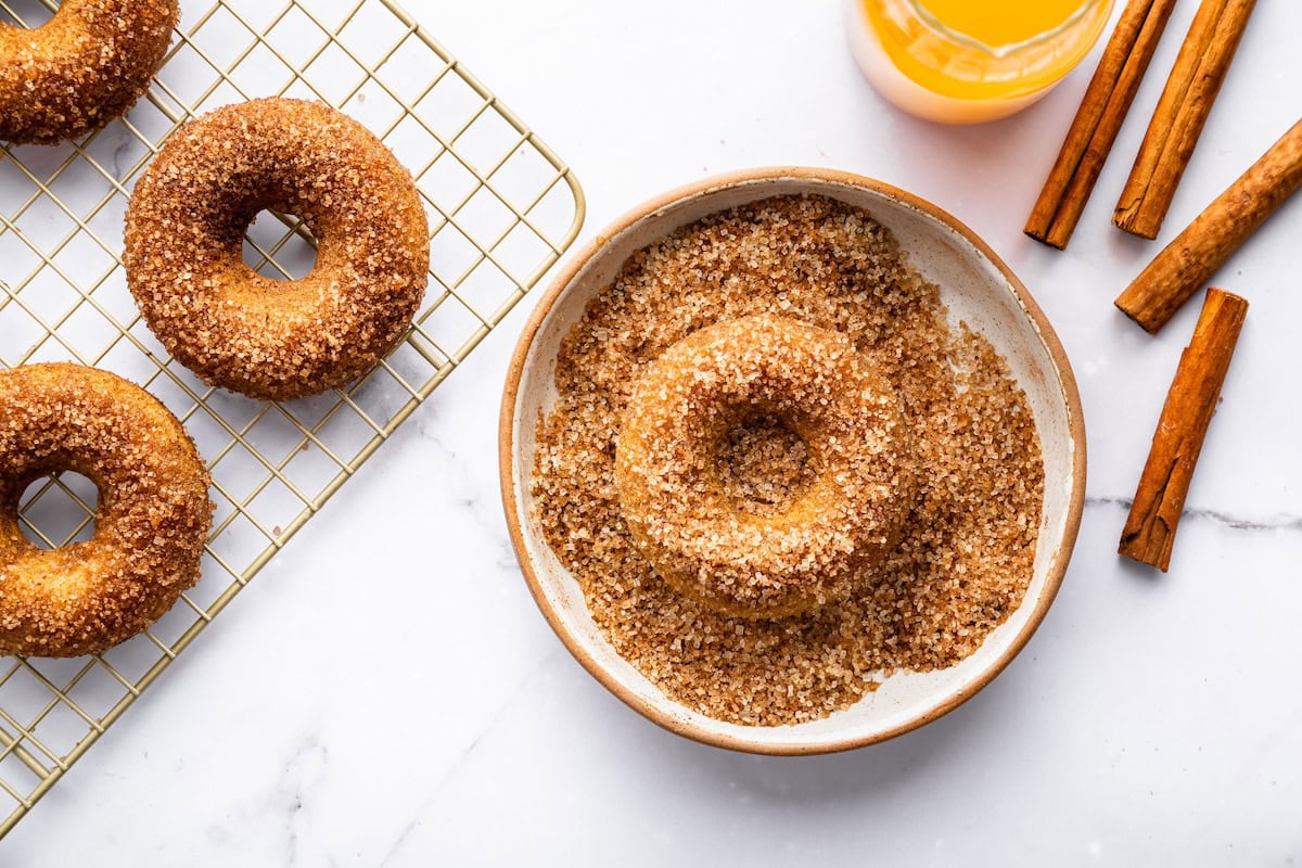 Apple cider donut being coated in a bowl of cinnamon sugar with more donuts cooling on a wire rack.
