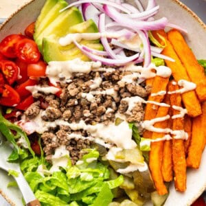 Assembled burger bowl with lettuce, tomato, onion, pickles, avocado, fries, and beef all drizzled with Big Mac sauce.