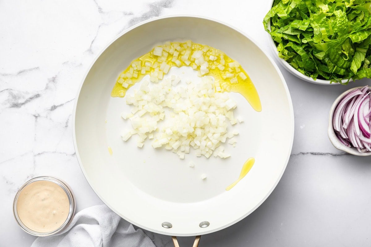 Chopped onions sautéing in oil in a white skillet.