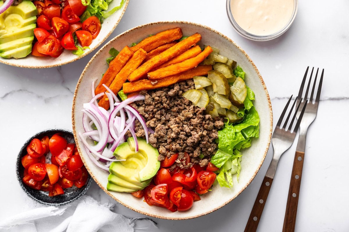 Burger bowl with ground beef, sweet potato fries, avocado, tomato, red onion, pickles, and lettuce.