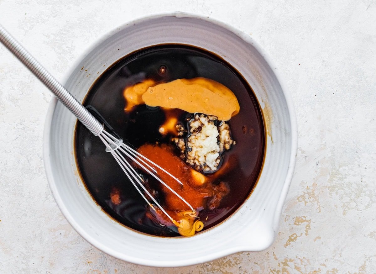 White mixing bowl with peanut sauce ingredients including soy sauce, peanut butter, and garlic.