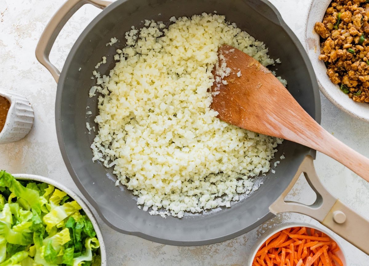 Cauliflower rice cooking in a skillet with a wooden spoon for lettuce wrap bowls.