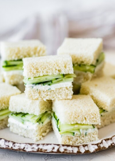 Pile of small cucumber tea sandwiches stacked on a white plate.