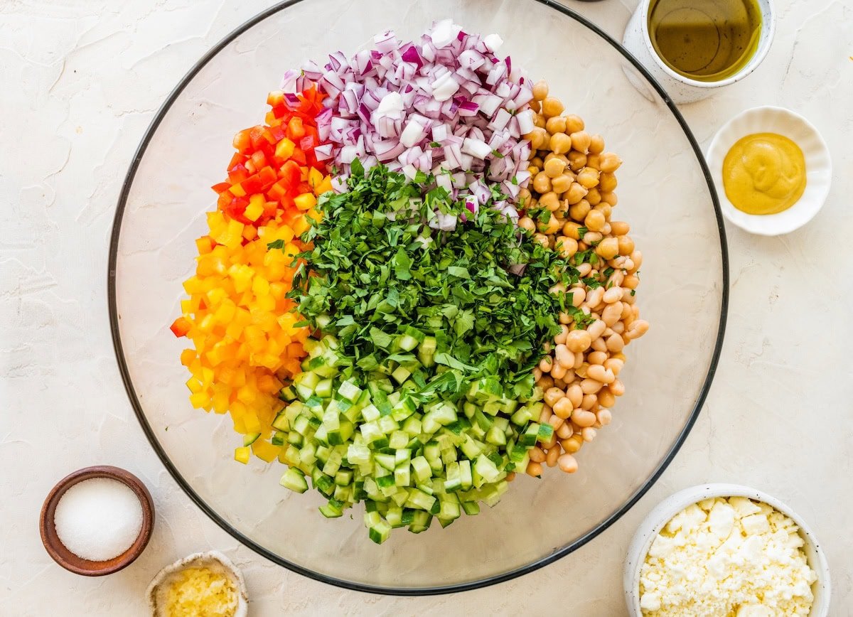 Large glass bowl with layers of diced red bell pepper, yellow bell pepper, red onion, chickpeas, white beans, chopped cucumber, and fresh parsley, surrounded by small bowls of dressing ingredients.