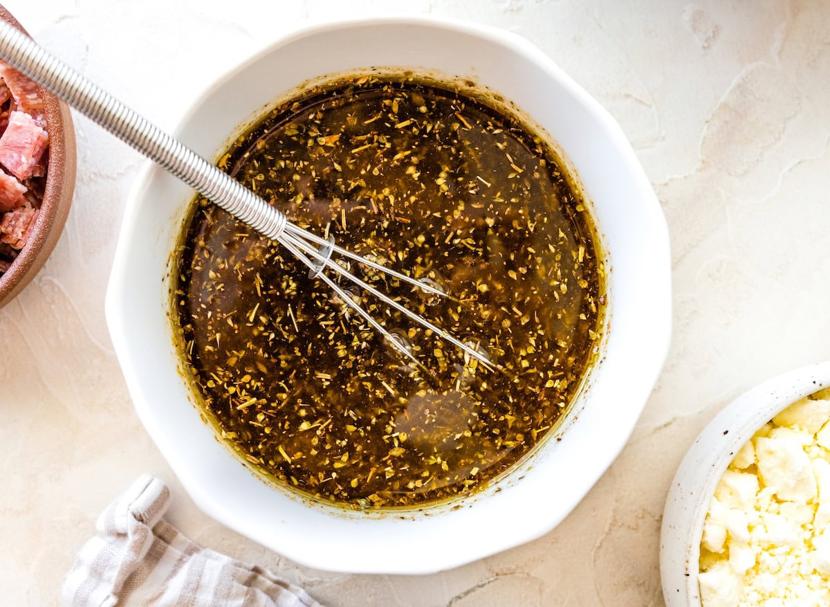 White mixing bowl of Italian herb vinaigrette with a whisk inside, next to a bowl of crumbled feta cheese.